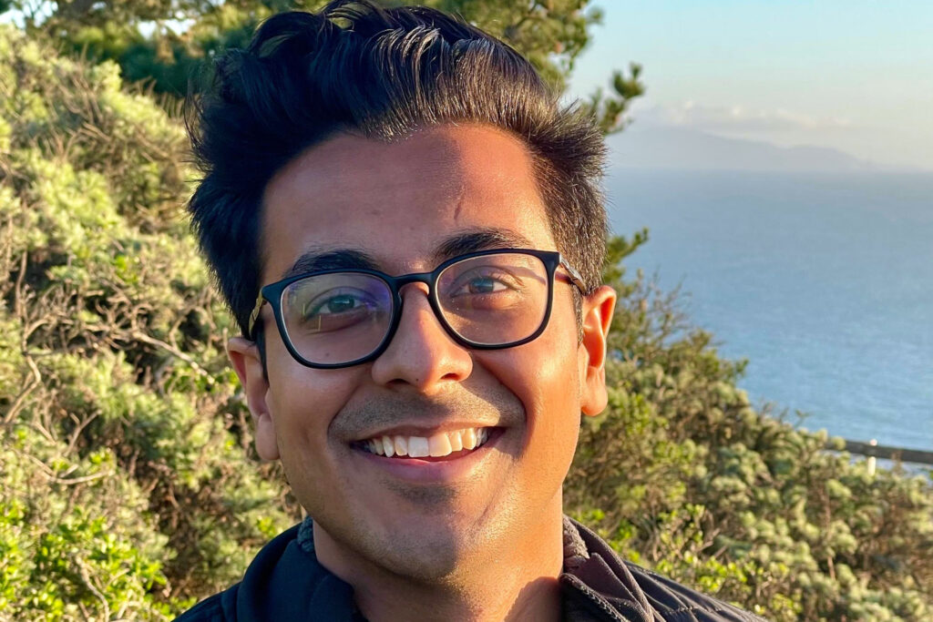 sunlit informal portrait of Berkeley alum Manu Meel, with green foliage and a large body of water in the background