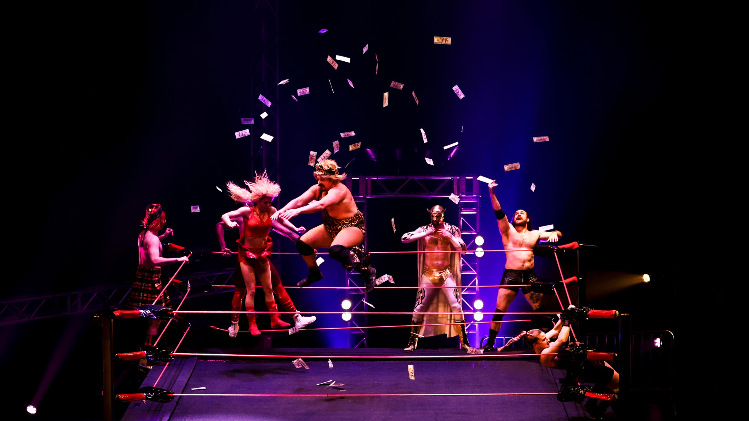 Seven acrobatic performers dressed like pro wrestlers perform a scene in a ring