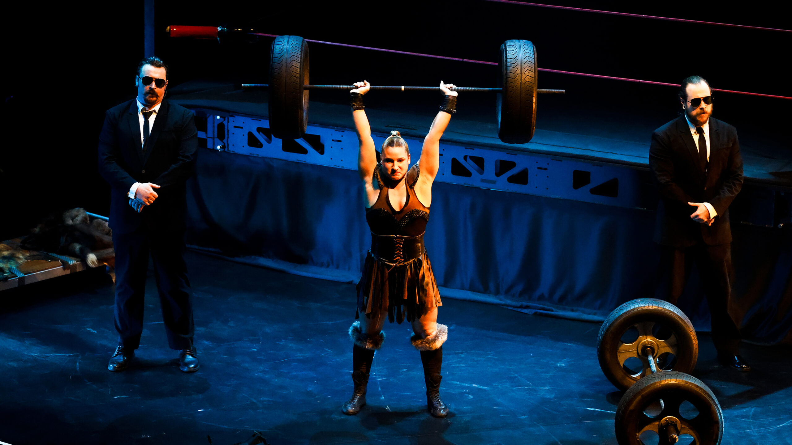 a cirque performer dressed as a pro wrestler holds up a pretend dumbbell while two men in suits and sunglasses stand on either side of her.