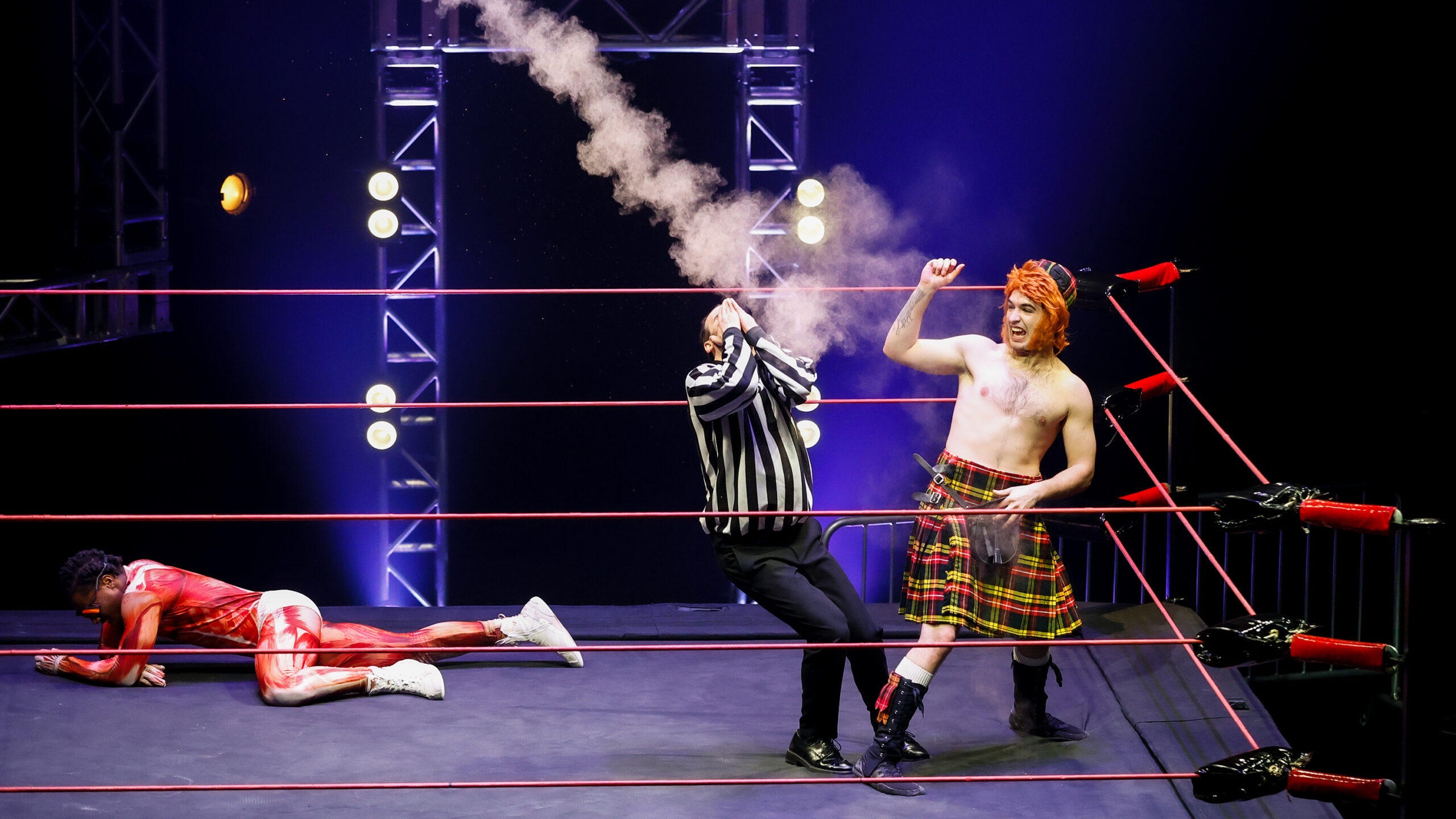 three cirque performers dressed like pro wrestlers in a ring. one dressed as a kilted Scotsman looks like he just threw a smoke bomb, the referee is holding their hands over their face and another is lying on the ground in defeat.