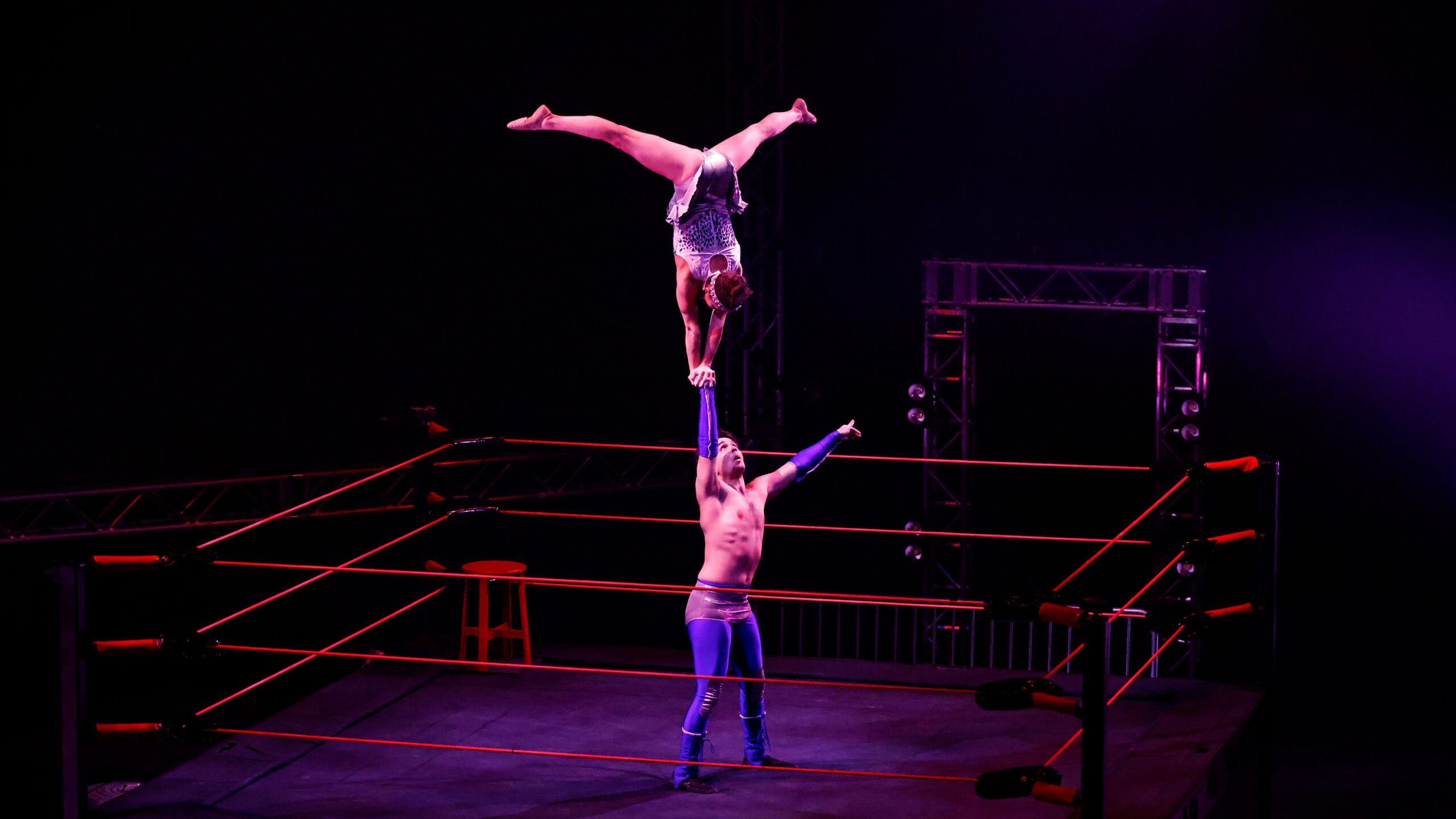 two acrobats dressed as pro wrestlers, one doing a handstand and splits on the other's hand outretched straight up