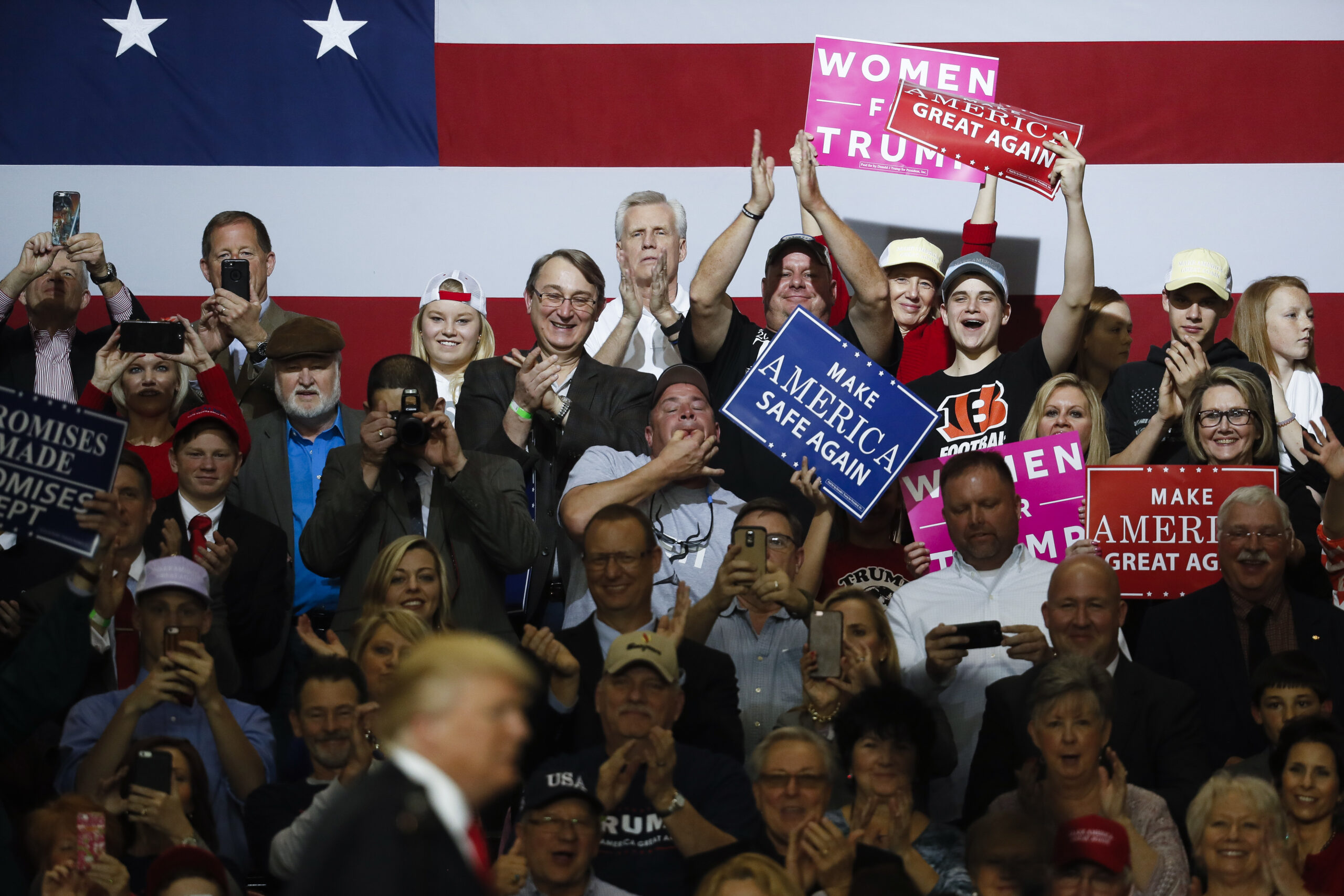 A crowd of supporters holding signs and wearing Trump-branded shirts cheer as Donald Trump, blurred in the foreground, walks onto a stage.