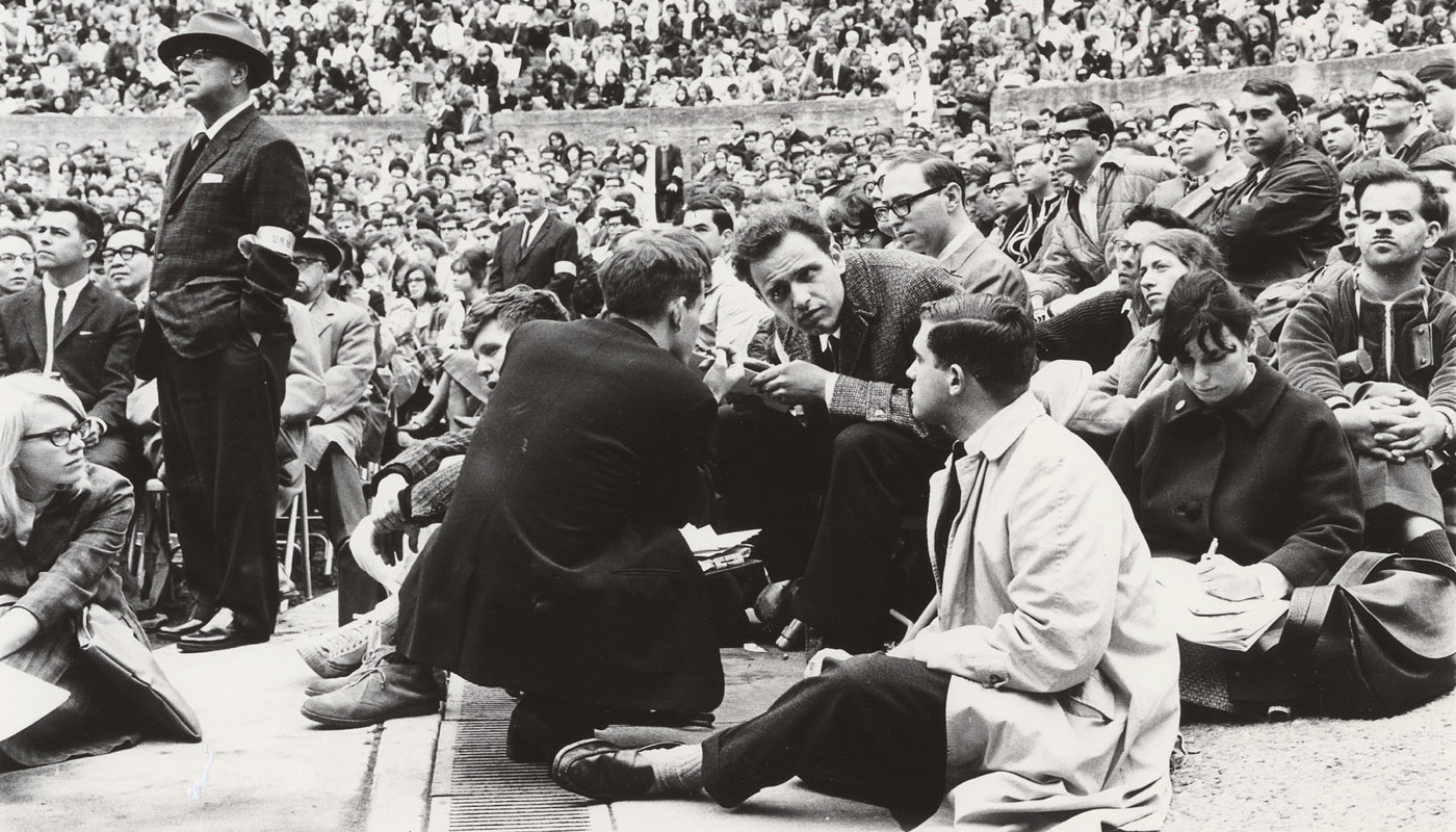 Black-and-white photograph showing activist Mario Savio in conversation with a group of people seated on the ground at the Greek Theater. The scene captures a large crowd of spectators in the background. Savio appears engaged in discussion, surrounded by people, some of whom are taking notes.