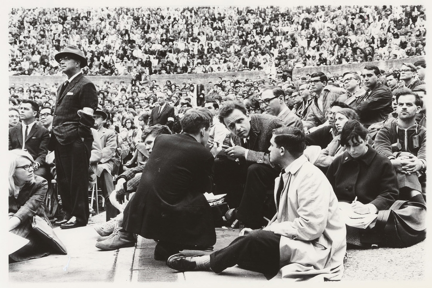 Black-and-white photograph showing activist Mario Savio in conversation with a group of people seated on the ground at the Greek Theater. The scene captures a large crowd of spectators in the background. Savio appears engaged in discussion, surrounded by people, some of whom are taking notes.