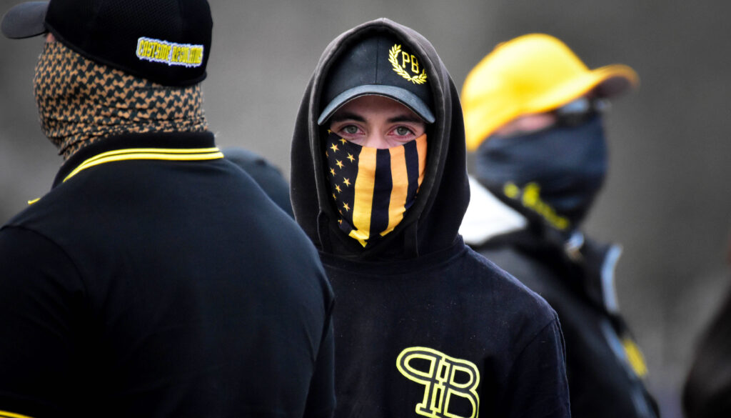 A young man attired in navy blue hoodie, baseball cap and a yellow-black US flag mask, with prominent Proud Boys insignias