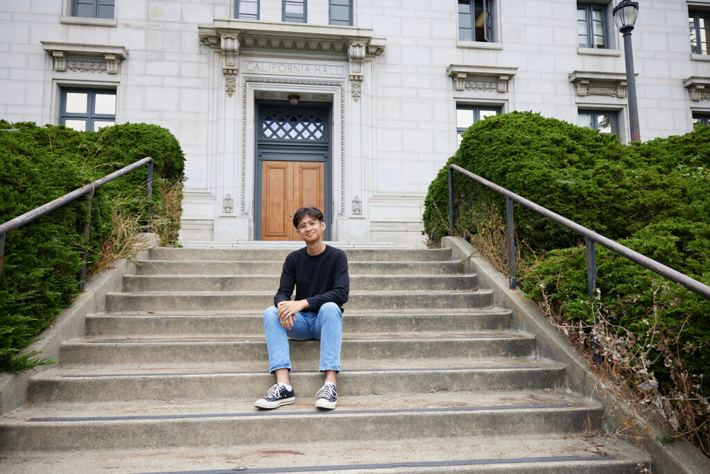 Student Aaron Occulto sits on the steps of California Hall. He is studying Burmese as part of the humanities majors he's taking. He is wearing jeans, a black T-shirt and black athletic shoes and smiling.