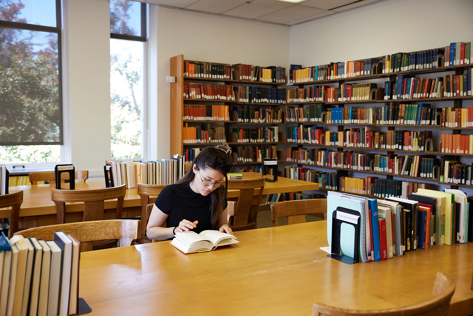 A student reads a book in a campus library and is sitting at a large wooden table.