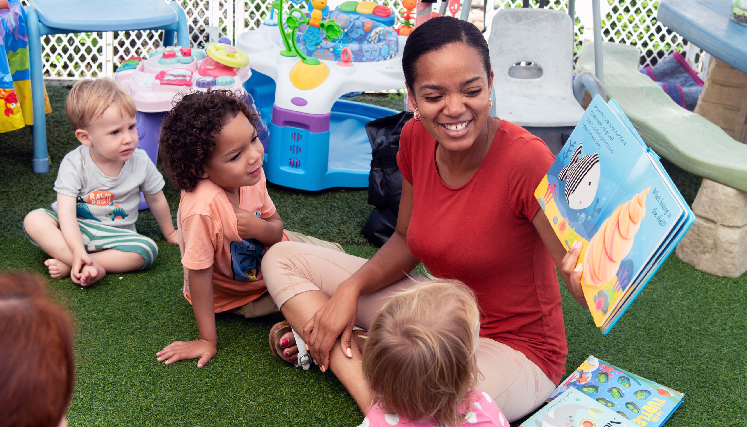 An early education teacher, holding an open books, sits cross-legged as she reads to a small group of toddlers.