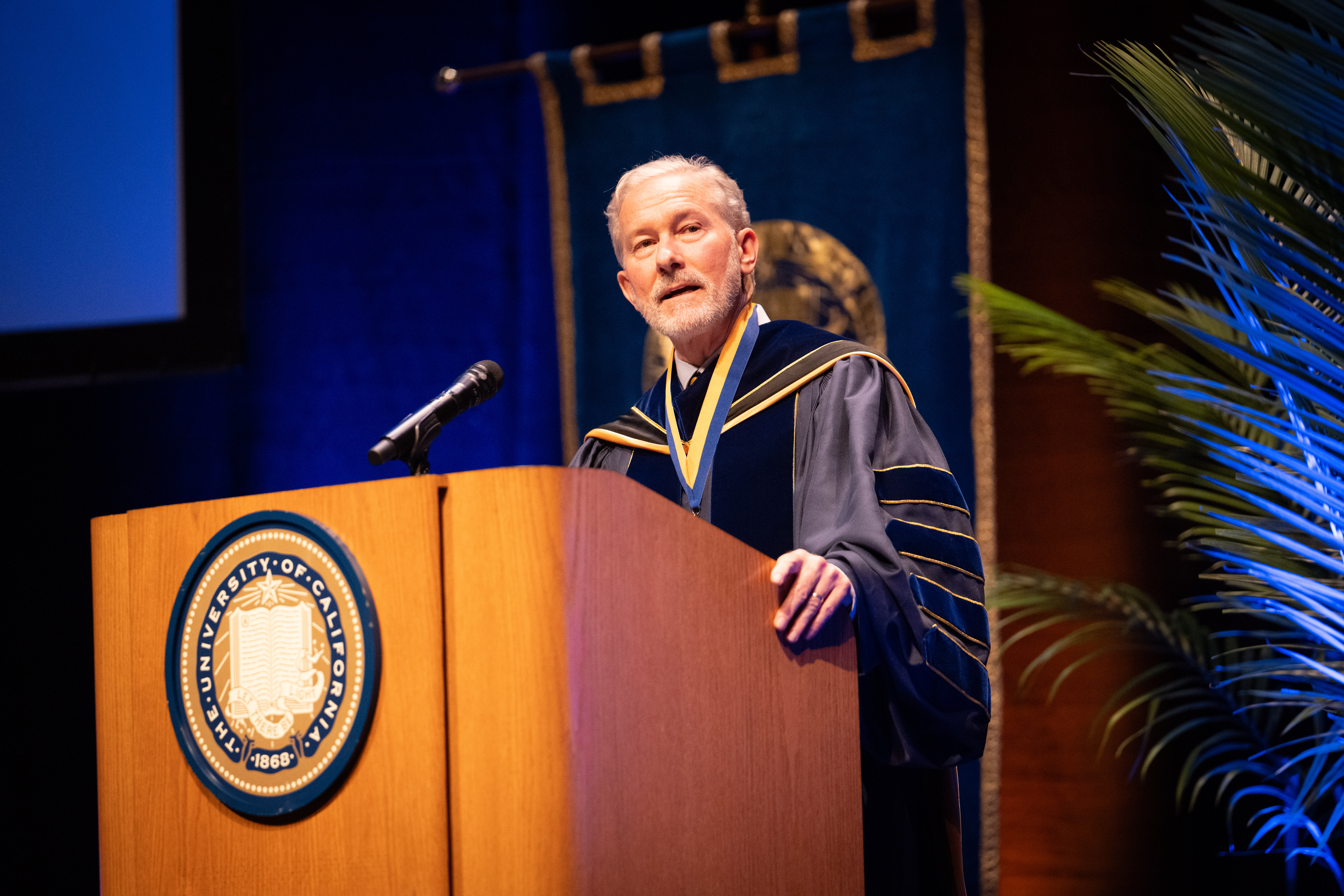 Chancellor Rich Lyons, standing at a podium and wearing his ceremonial robes, speaks into a microphone during his inauguration ceremony.