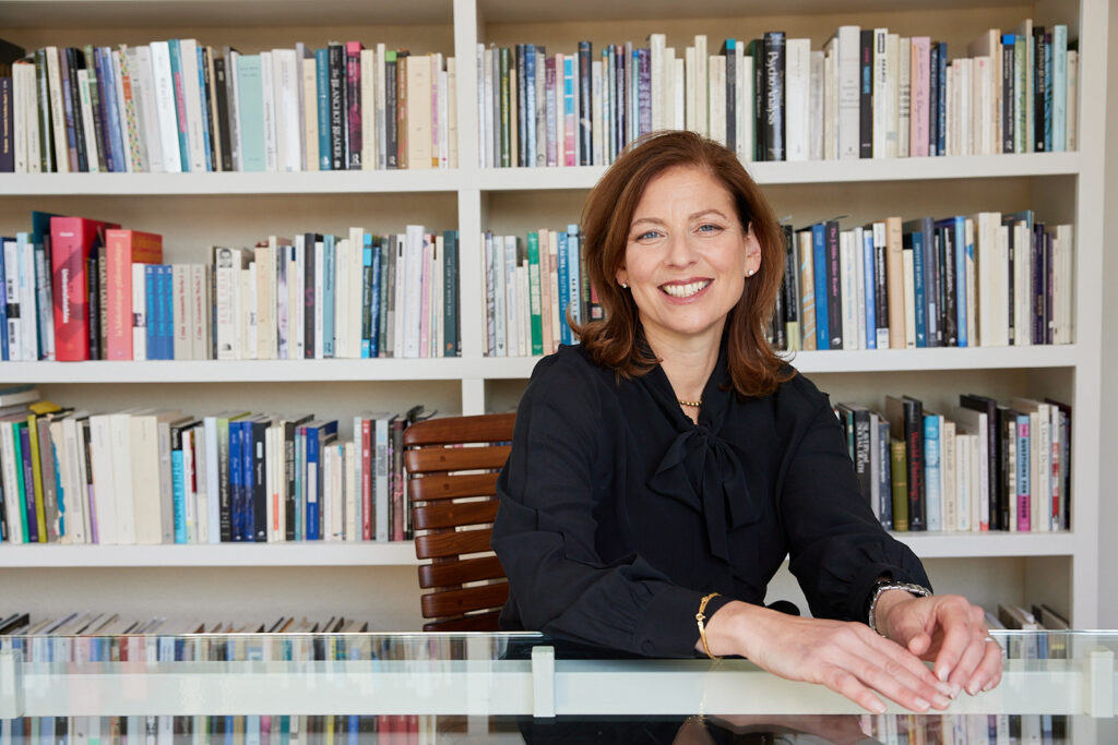 Sara Guyer, dean of the Division of Arts and Humanities in the College of Arts and Sciences, sits at a desk smiling. There is a bookshelf behind her with hundreds of volumes.