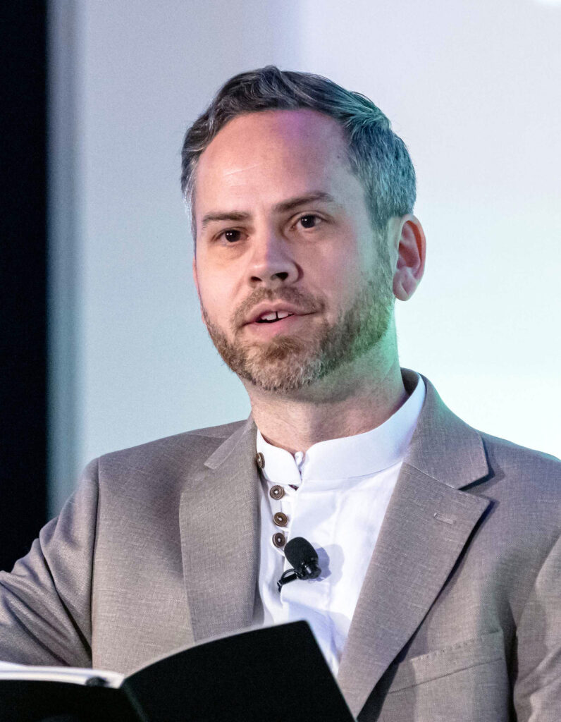 casual headshot of Joshua Clark, reading from a black notebook at a conference