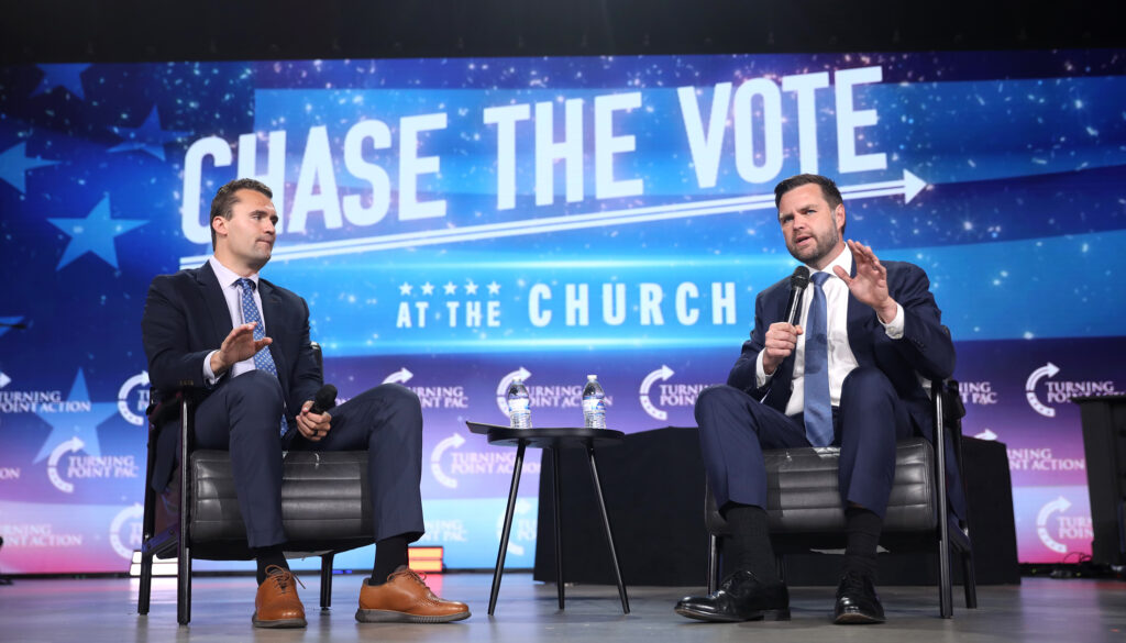 J.D. Vance and Charlie Kirk giving a talk on stage at an event with "Chase the vote at the church" projected on a huge screen behind them