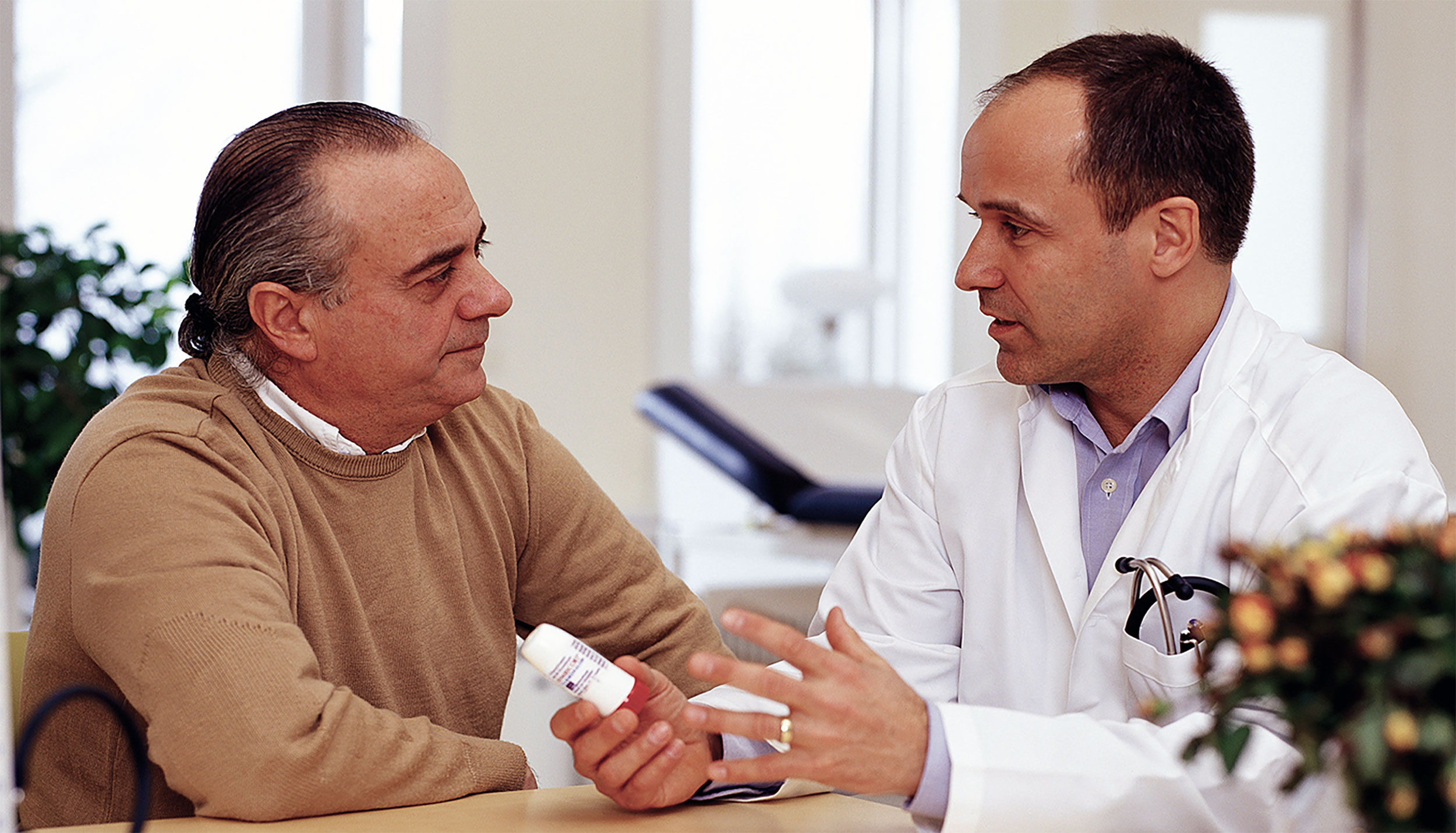 an older man, at left, confers in a medical office with a doctor (at right) who is holding out a prescription vial.