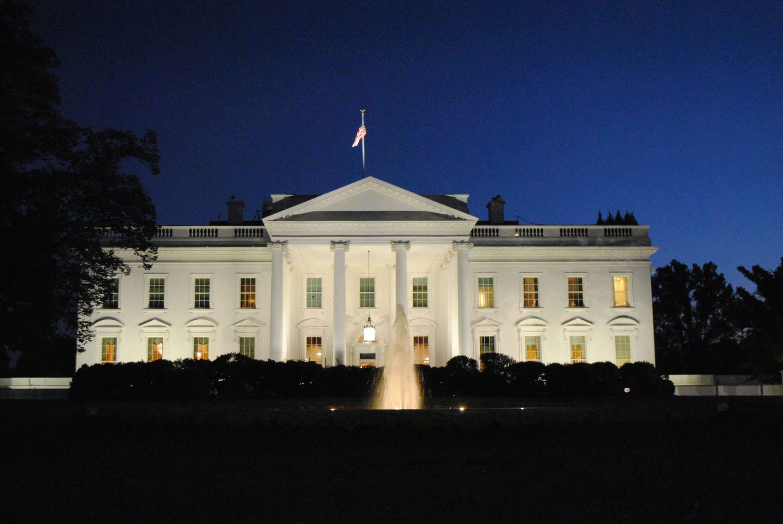 The White House in Washington D.C. at night, illuminated with lights and with a fountain in the foreground shooting water into the air.