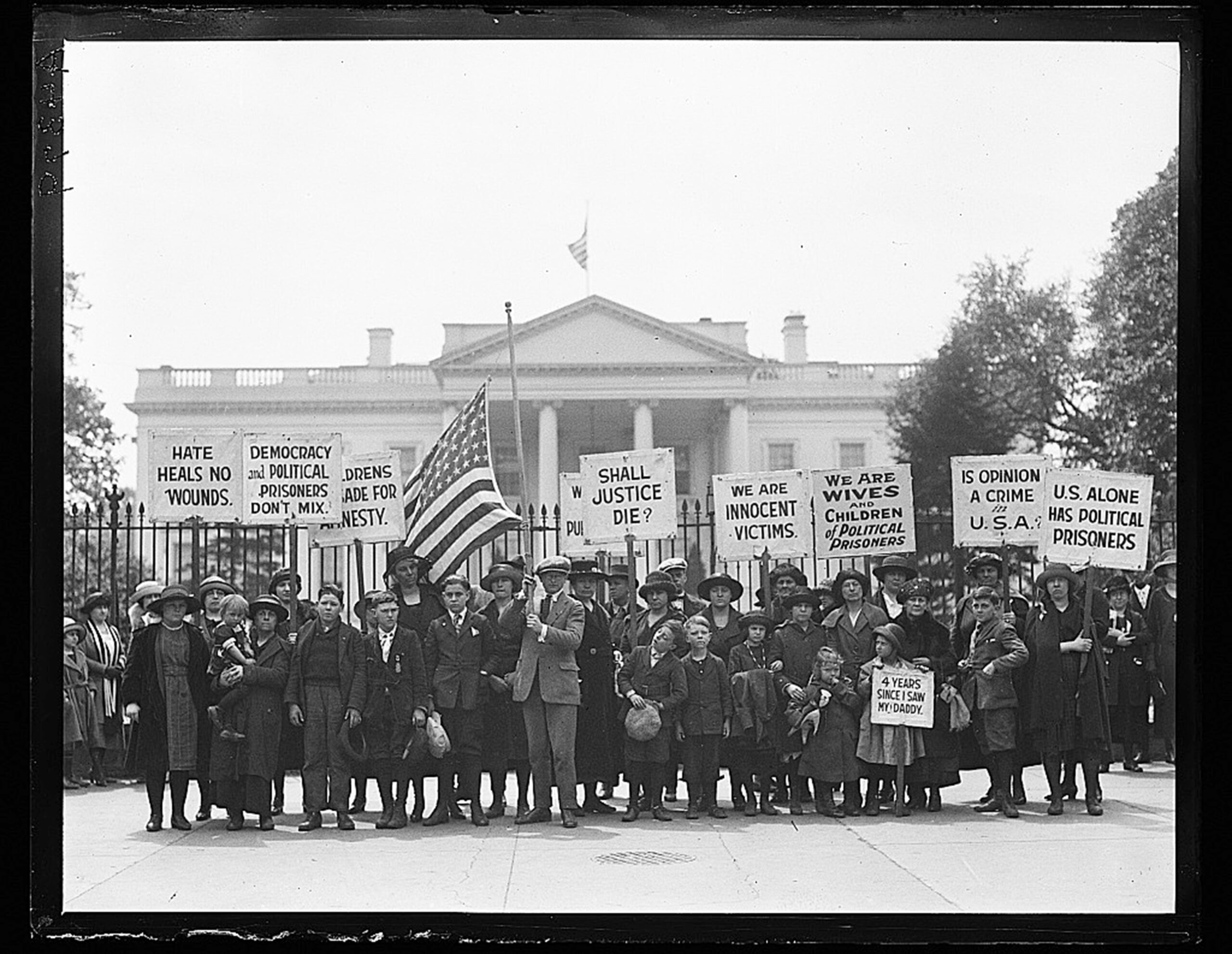 In a black and white photograph, a group of people, including children, stand in front of the White House holding signs that say 