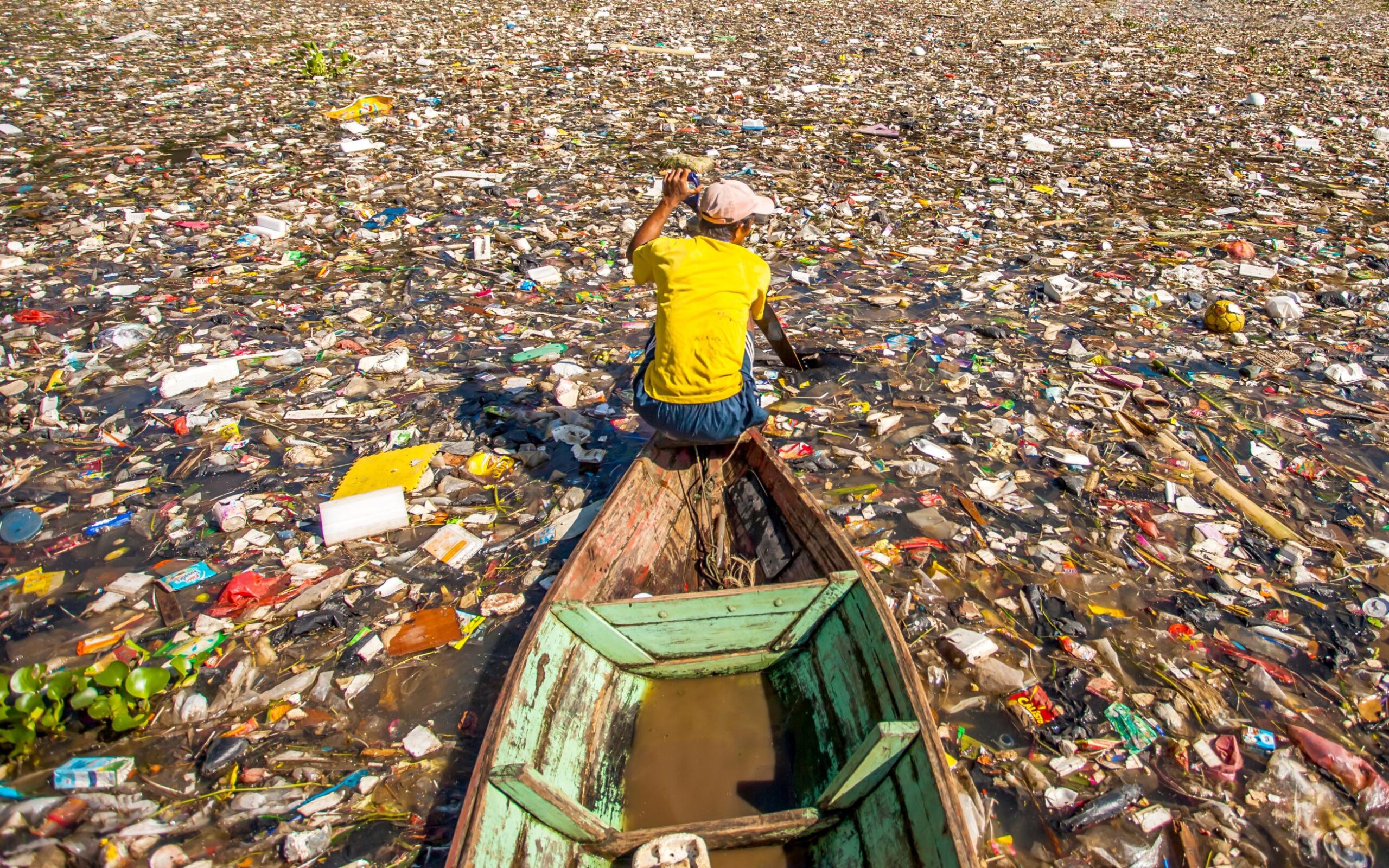 A person rows a canoe through water that is filled with floating garbage.