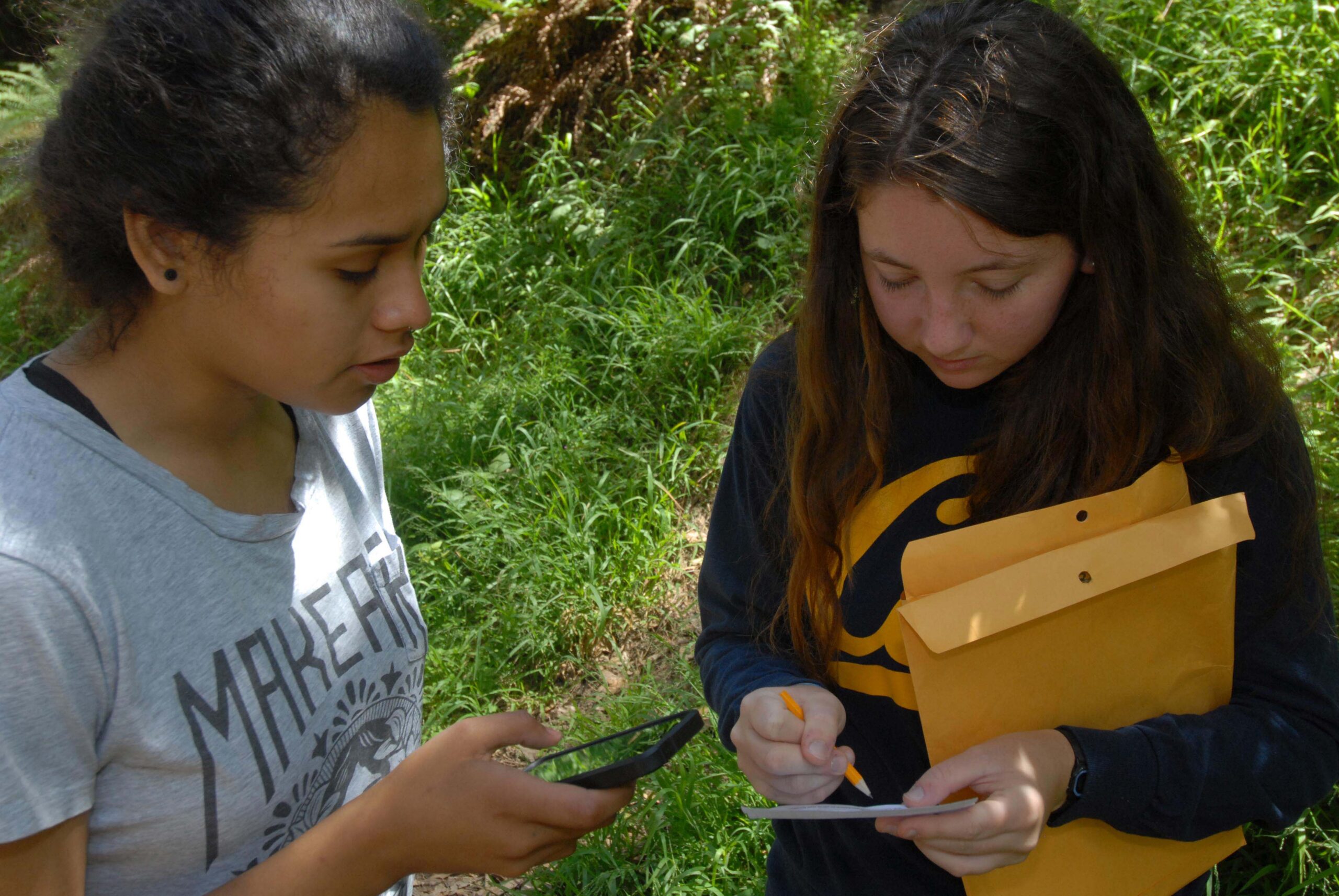 Two people stand next to one another in a forested area. One person is reading off of their phone while the other is writing something on a small piece of paper.