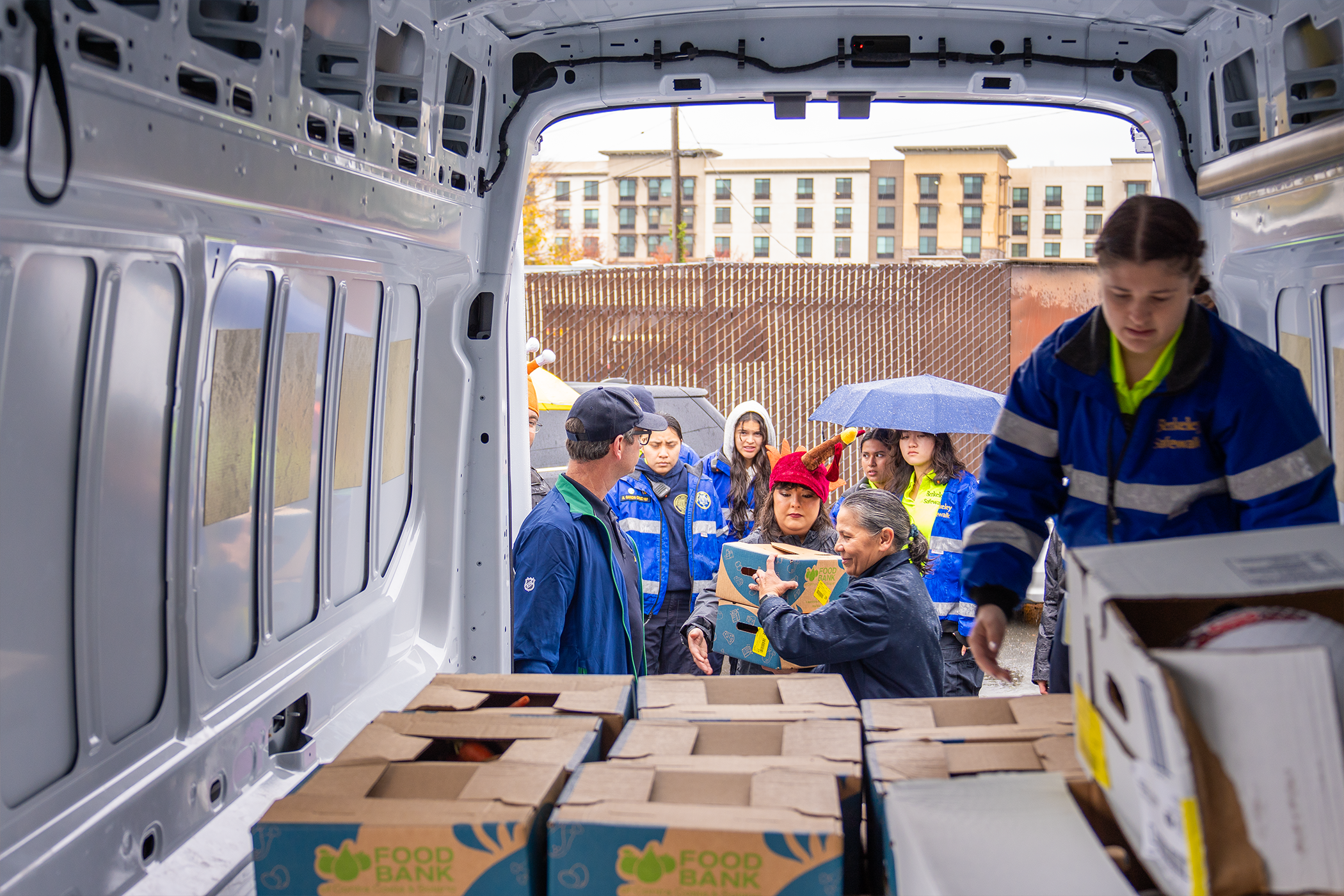 A person wearing a Berkeley Safewalk jacket reaches toward boxes in the back of a cargo van, while another person standing outside the van hands boxes to others.