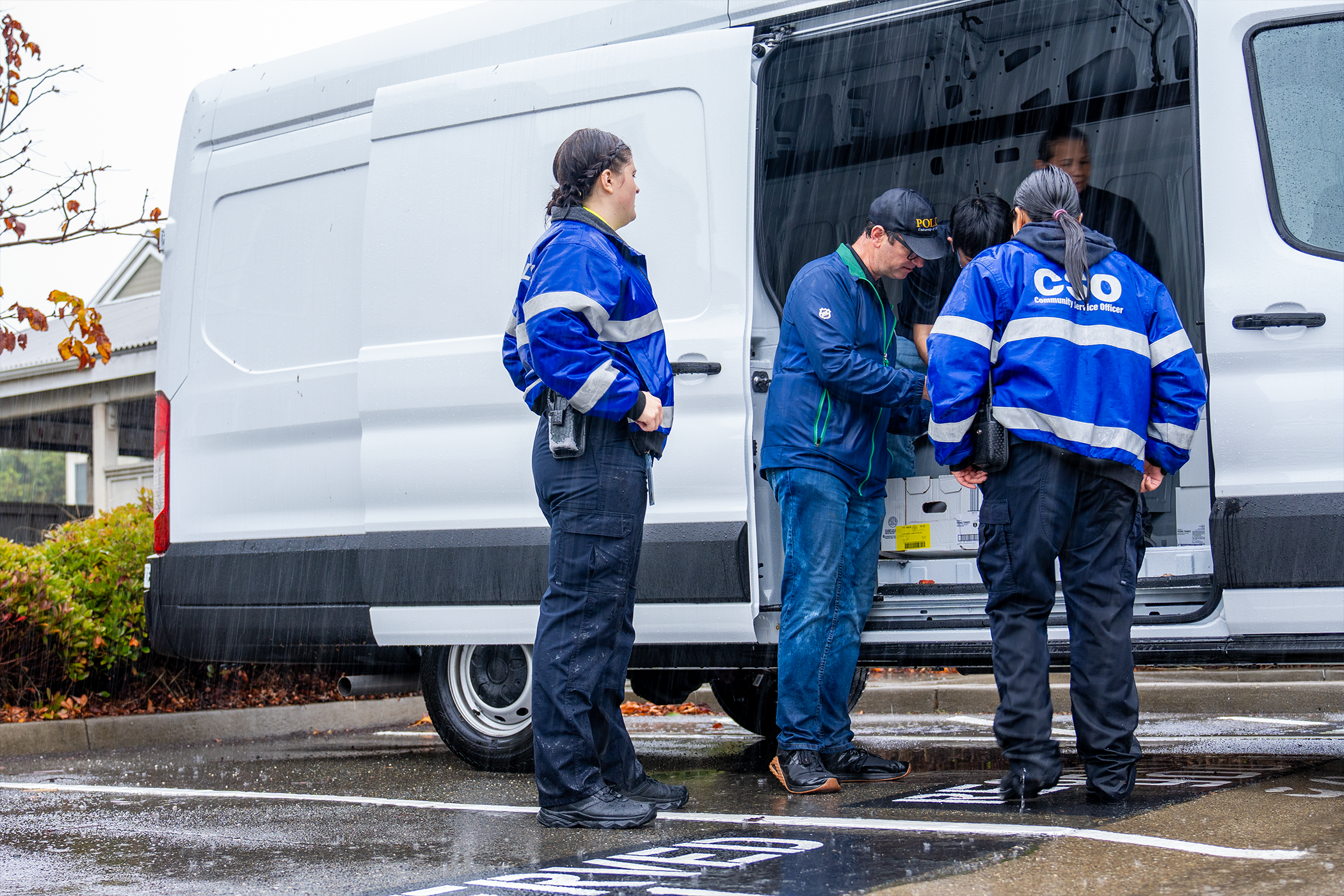 Five people stand near a white van, consulting a list of addresses to for meal deliveries while rain streaks down in the foreground