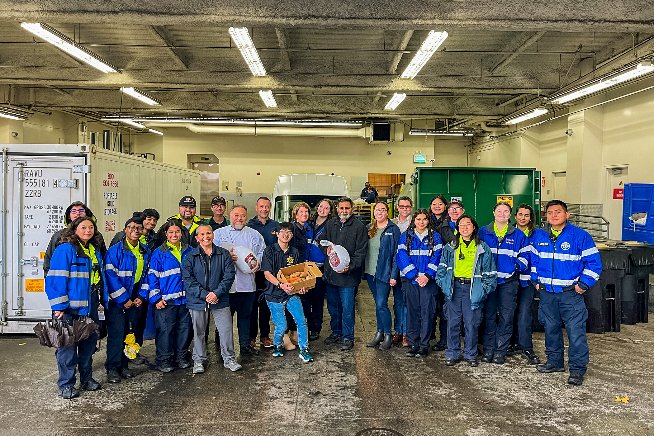 Approximately two-dozen smiling people stand posed for a photo in a loading bay, near the van that delivered the meals to University Village. Three of the people are holding frozen turkeys and produce.