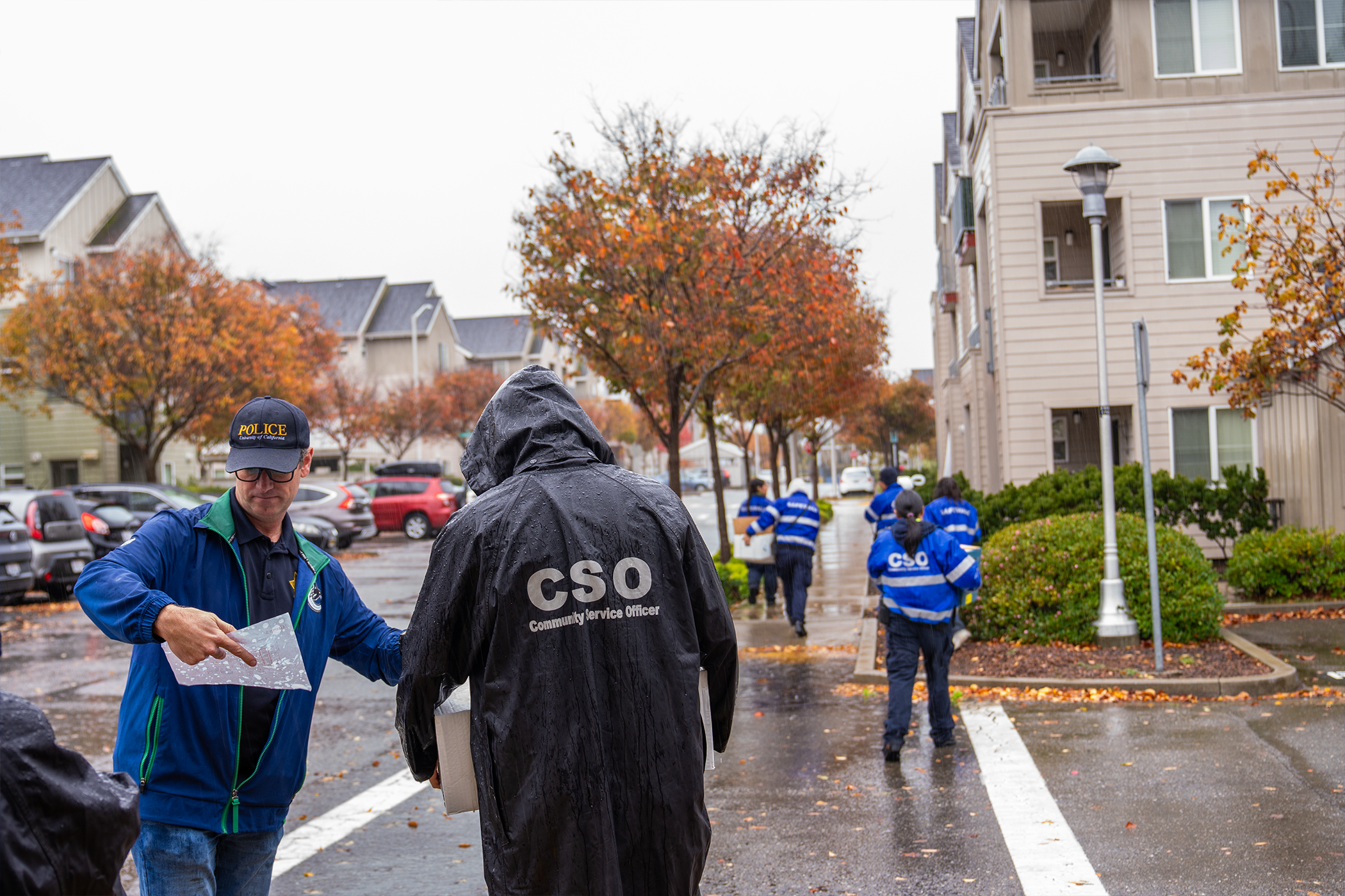 A person in the lower right reviews a piece of paper with addresses while 6 people carrying boxes of food walk toward a building