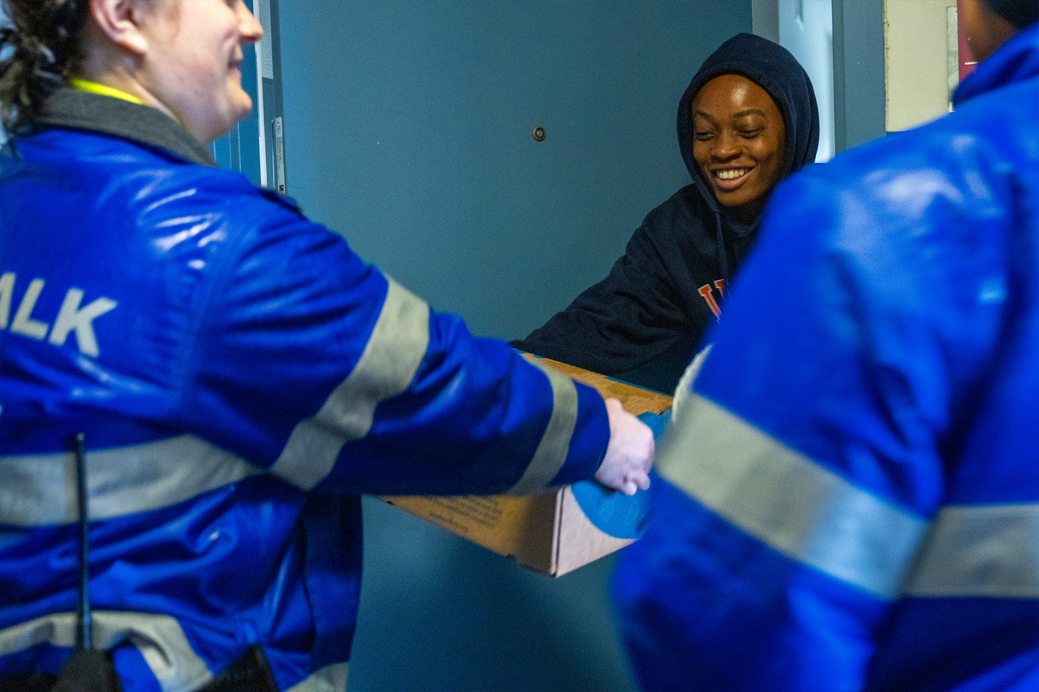 A person wearing a sweatshirt reaches for a box of produce being handed over by a woman wearing a blue Safewalk jacket