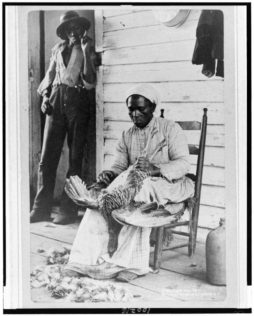 A Black woman with her hair covered sits in a chair and plucks a turkey as a Black man watches and smokes in the black-and-white photograph. There are feathers on the ground.