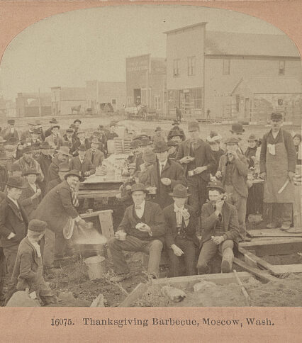 A sepia photograph of men in suits and hats sitting and standing around tables. Some are eating turkey wings, and one appears to be doing some food preparation.