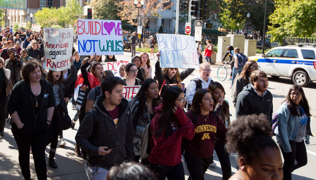 A peaceful student protest featuring placards reading "Build Love Not Walls" and "1st gen American against Racism"