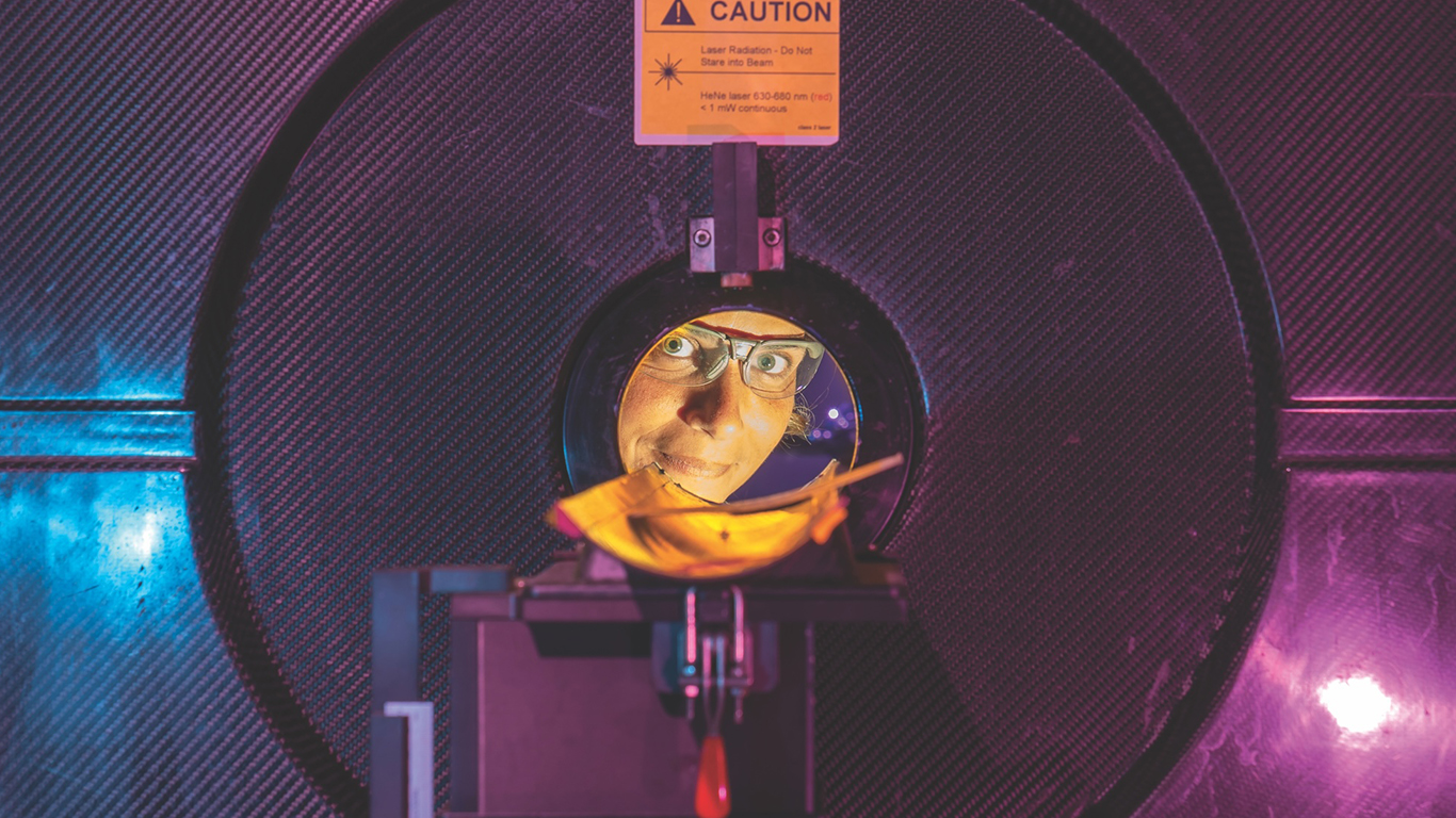 A person wearing safety glasses is seen looking through the porthole of a large experimental chamber that is bathed in purple light.