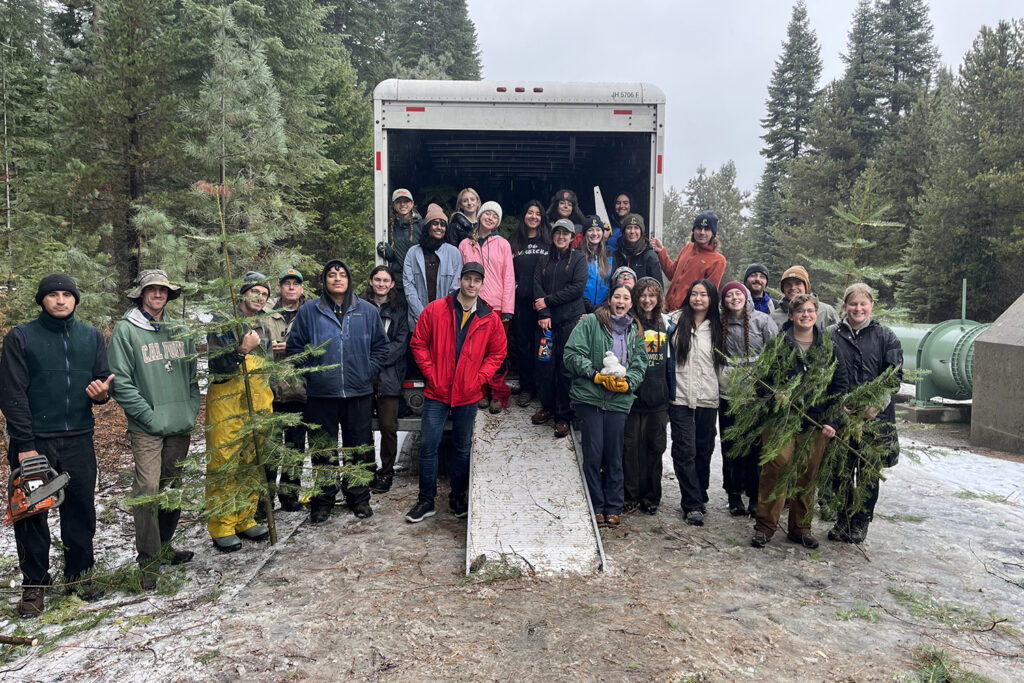 A group of about 20 students in the Sierra pose next to the UHaul truck that they're filling with cut Christmas trees for their annual sale on campus.