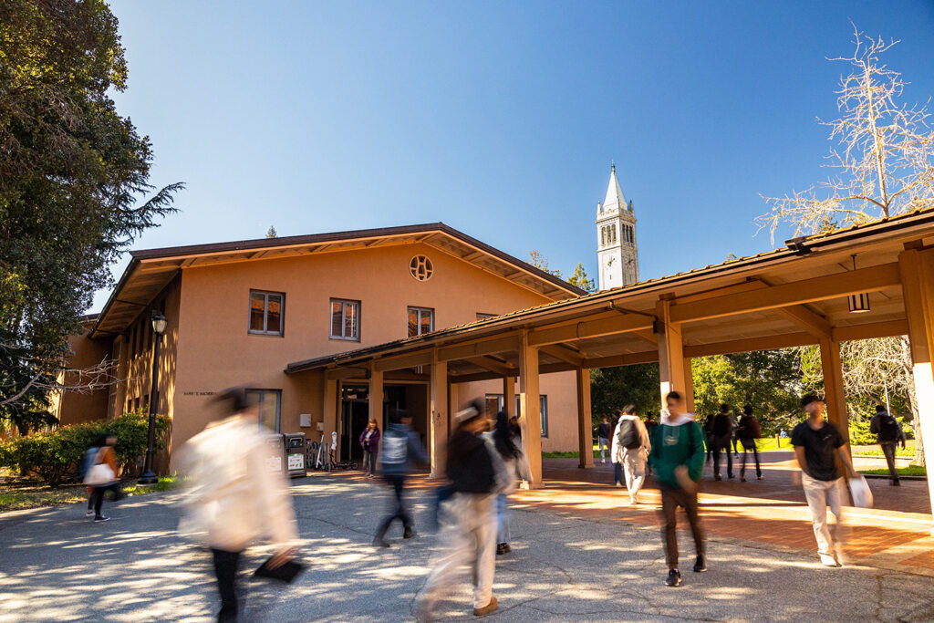 The outside of Morrison Hall, home to the music department, shows students walking past with the top of the Campanile in the background, over the rooftops.