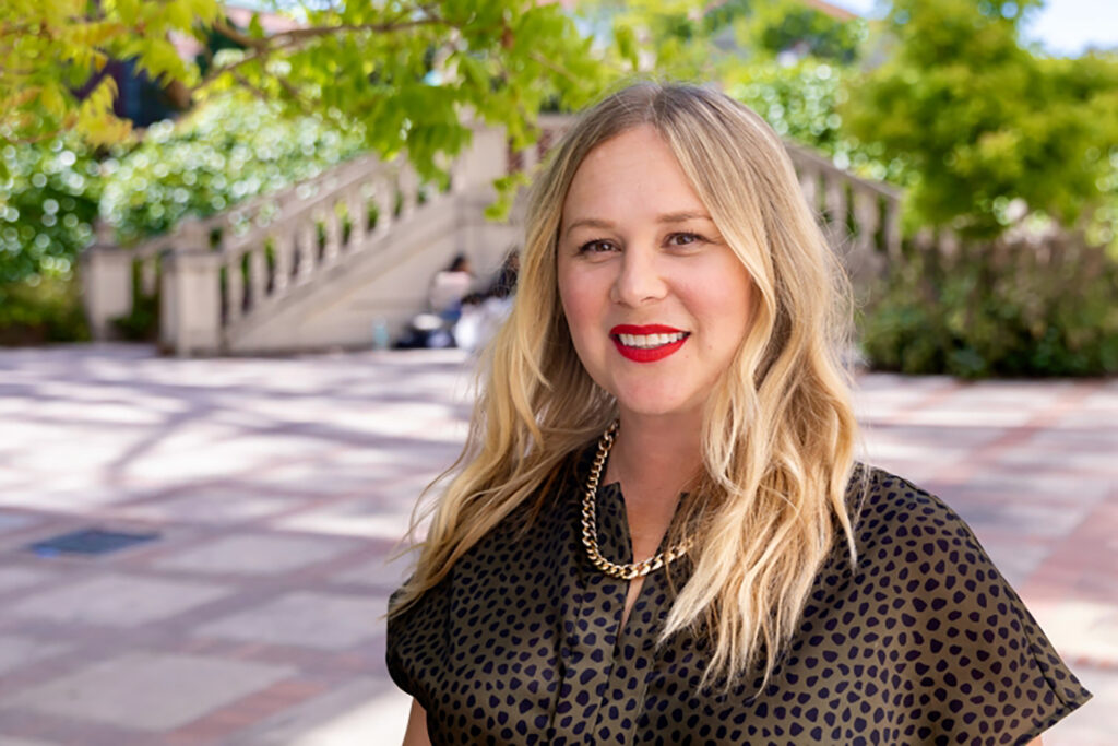 Rhiannon Welch, the Giovanni and Ruth Elizabeth Cechetti Chair of Italian Literature, smiles at the camera in front of a stairway near Stephens Hall. She has long blonde hair an amimal print top on, and a thick gold chain as a necklace.