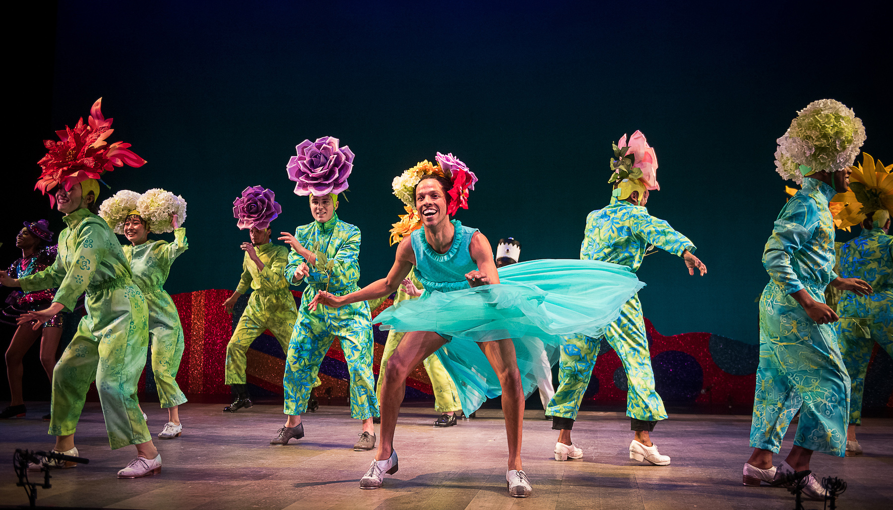 about a dozen dancers dressed in bright costumes with giant fabric flowers on their heads perform on stage in the nutcracker suite.