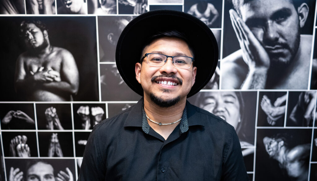 Brandon Sánchez Mejia stands in front of his photography exhibit, photos of men's faces and bodies in various poses to reflect vulnerability