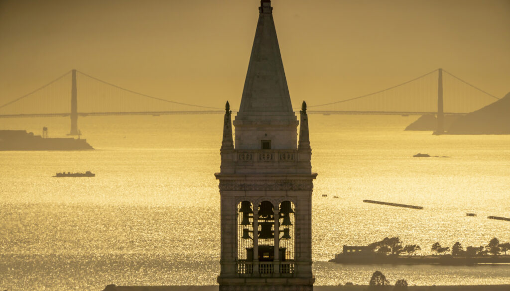 a bell tower stretches into the sky with the San Francisco Bay and a bridge in the background. The light is golden in color, showing the sun setting.