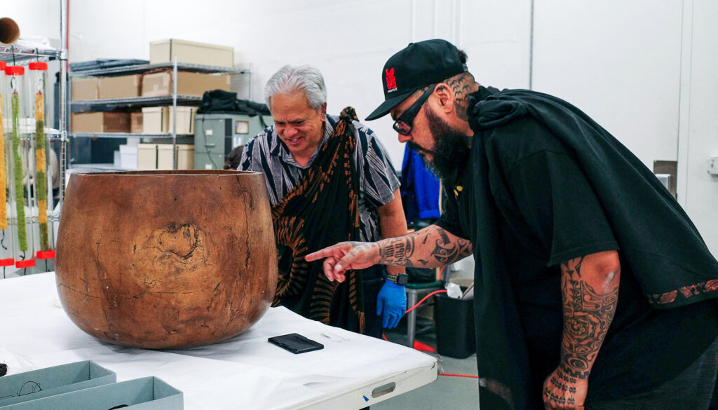 Halealoha Ayau and Mana Cáceres bend to look and gesture toward a large tan-colored 'umeke nui, a type of storage container that has a woodgrain-style finish.