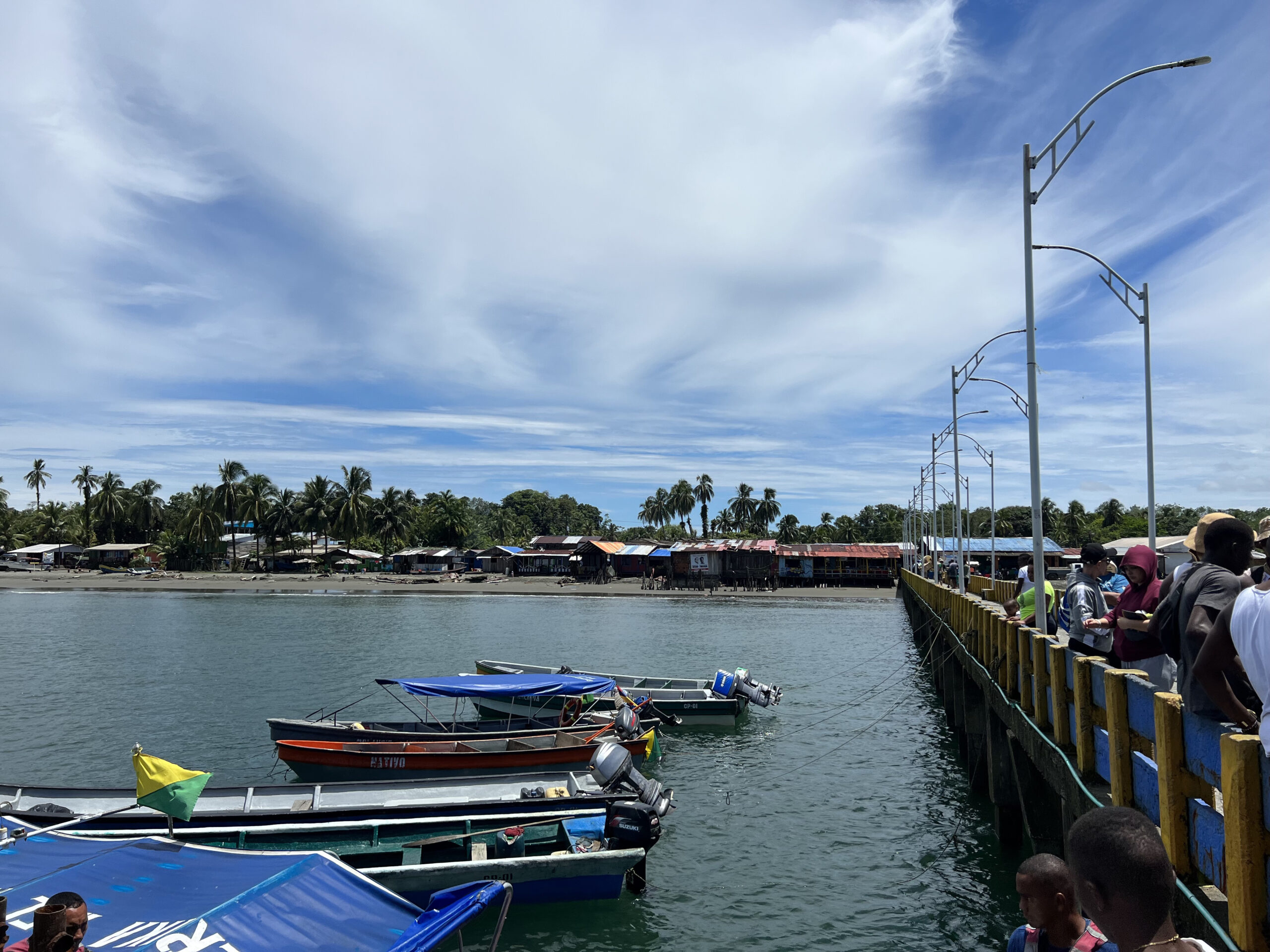 small boats bobbing in the water alongside a pier, with wispy clouds against a blue sky