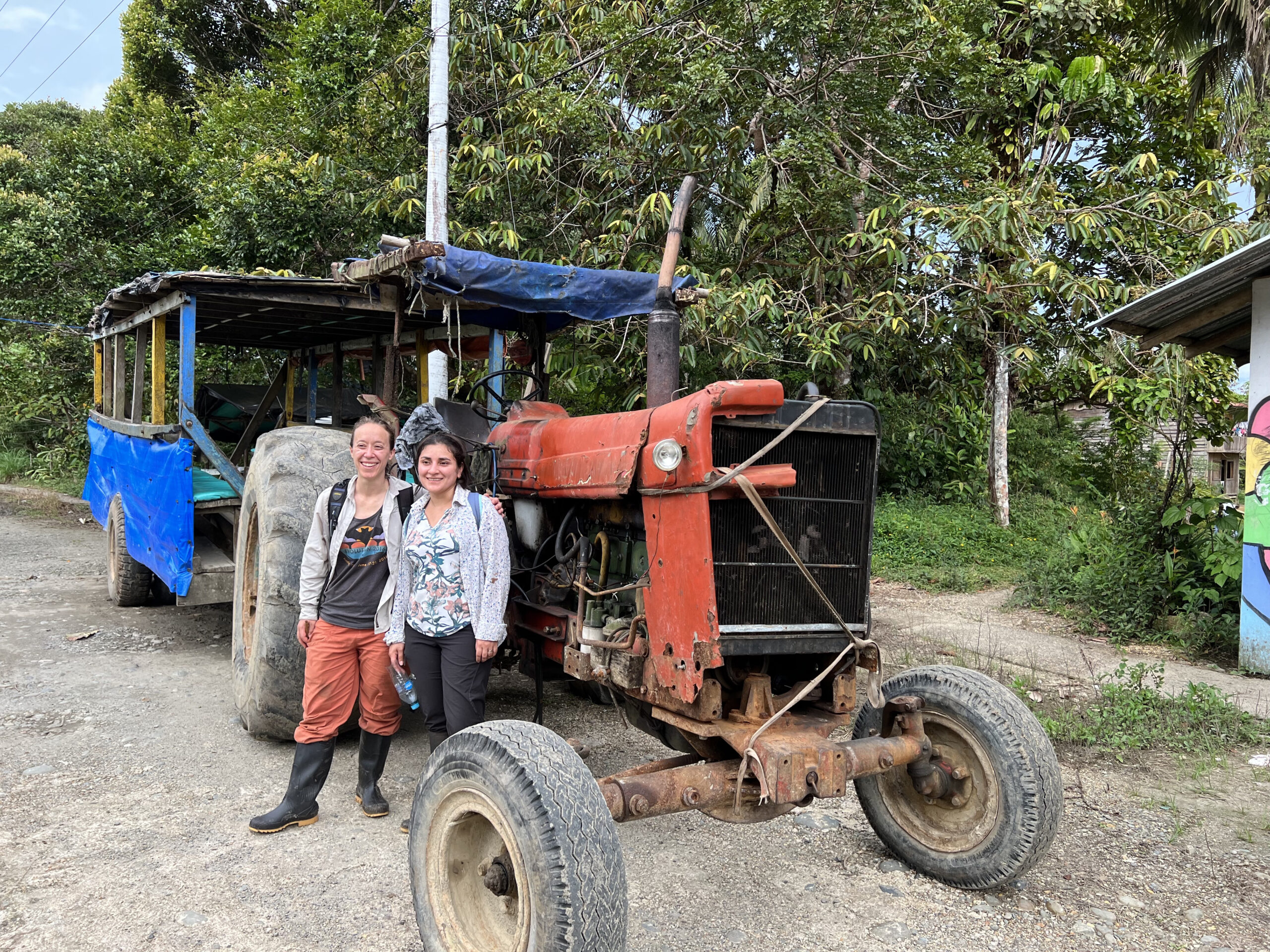 two women standing next to a rusty orange tractor with an attached open-air carriage covered in a blue tarp