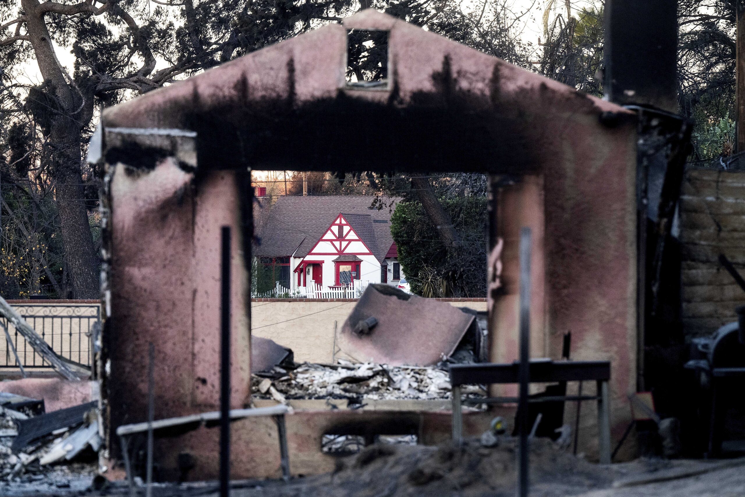 A charred building, surrounded by debris and ash with a view through the structure toward a white-painted home that is intact and undamaged by fire.