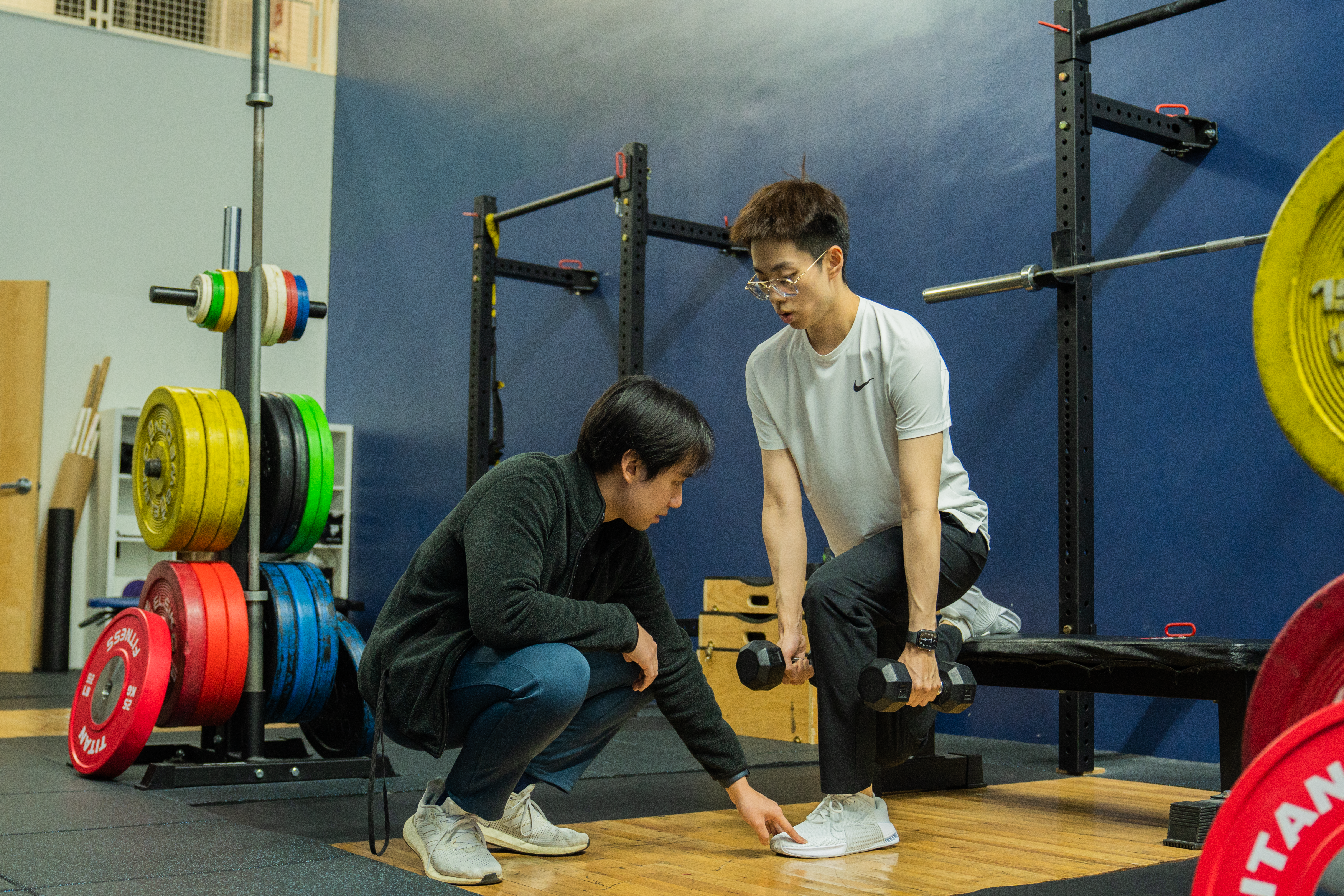 a trainer wearing dark clothing kneels and directs a student who is doing a squat with dumbbells