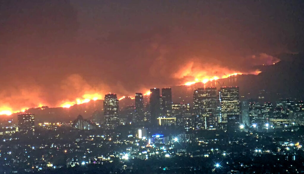 The Palisades Fire burns across a ridge behind the skyline of downtown Los Angeles