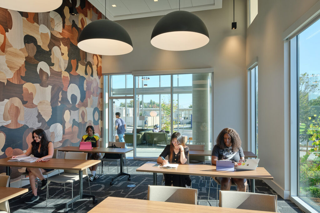 A view of a study area in xučyun ruwway student housing. Students are sitting at tables with books and laptops. The is a large mural on one of the walls that features the faces of people of many races, as well as large windows and doors that let the sunshine in.