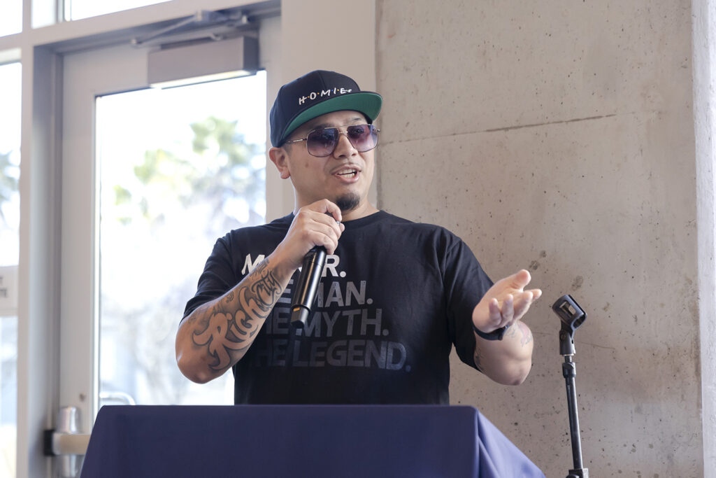 Wearing a cap, T-shirt and sunglassess, Ph.D. candidate Robin Lopez, who also is the mayor of Albany, holds a microphone and speaks at a celebration for xučyun ruwway, Berkeley's newest housing for graduate students.