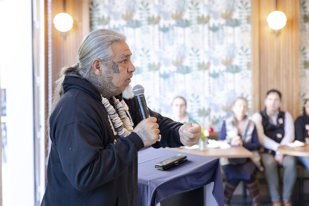 A Native American sings and strikes a clapper stick, giving a prayer at an event to mark the opening of UC Berkeley's newest housing complex, xučyun ruwway, in Albany. He has a long grey ponytail and a strong of large shells around his neck.