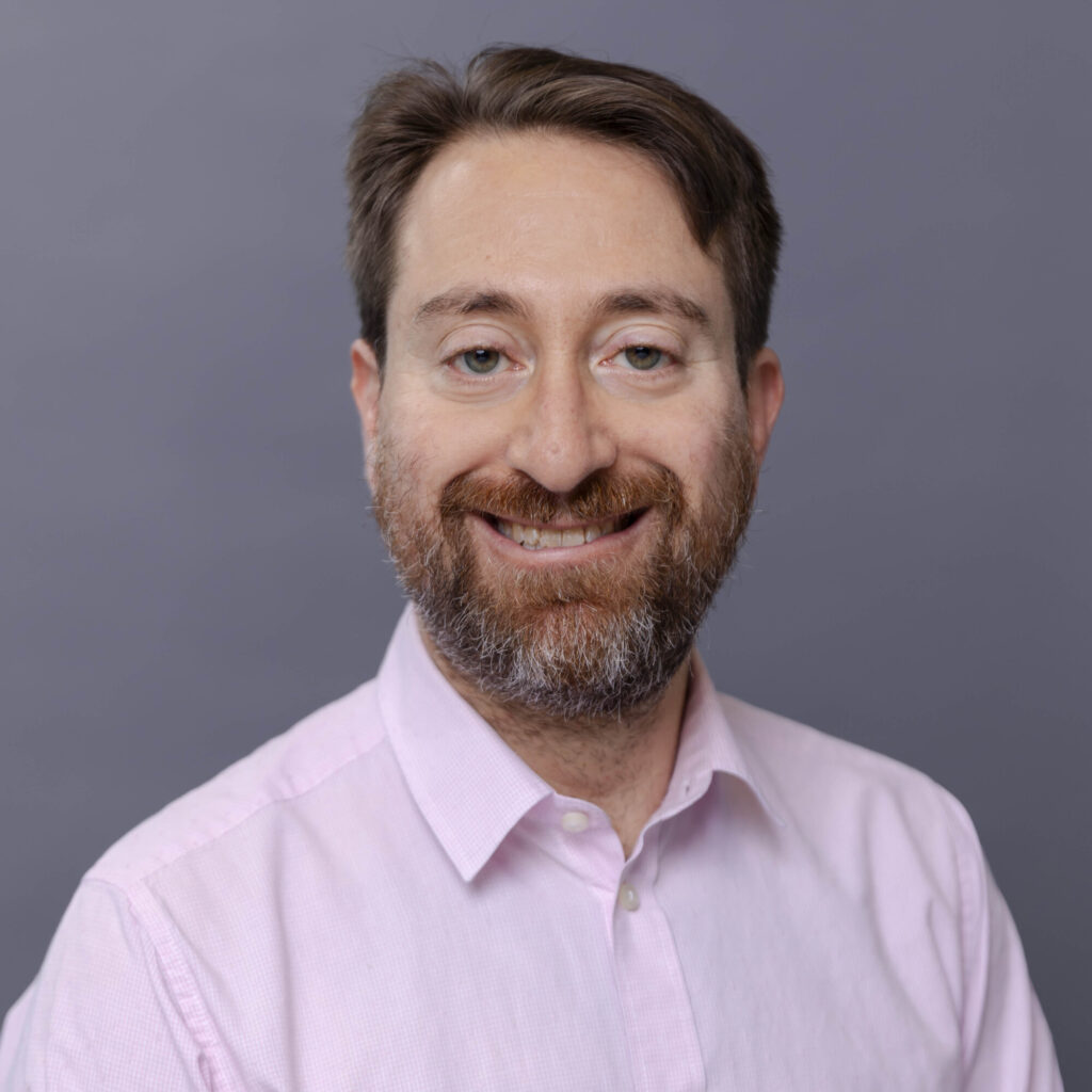 A headshot of a bearded man wearing a pale pink button-up.