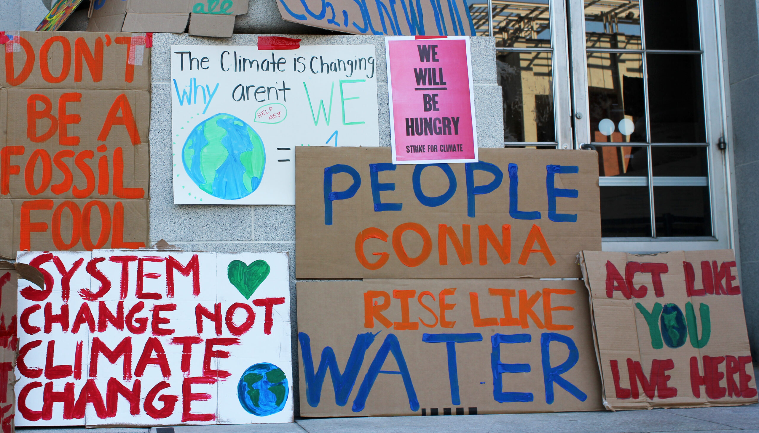 A collection of posters about climate change and the need to take policy action are posted on the steps of a public building. The posters have various messages: 