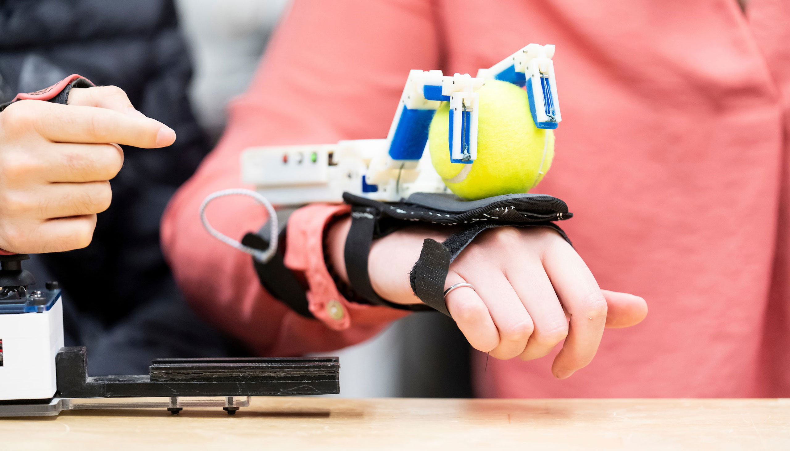 A person is pictured holding their arm above a table. A device is strapped to the back of the person's wrist and hand, and the device is grasping a yellow tennis ball.