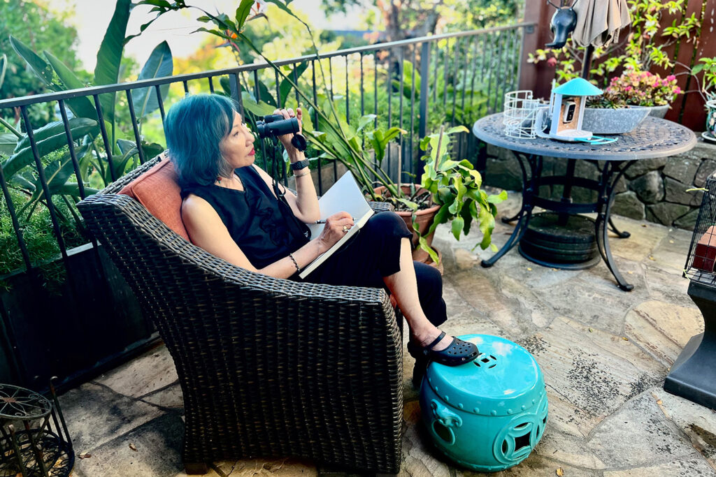 Author Amy Tan sits in a wicker chair on her backyard deck with binoculars and a notebook that she's filling with entries about birds.