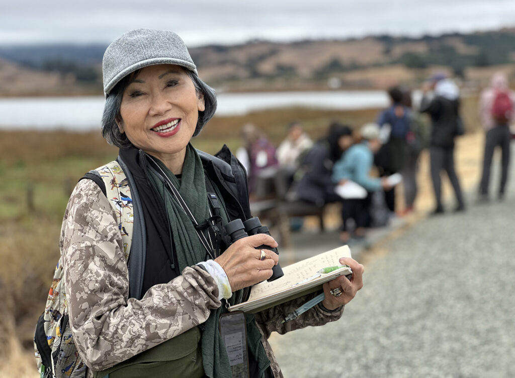 Author Amy Tan is outdoors, wearing warm clothes, binoculars and holding a sketch book where she makes drawings and notes. 